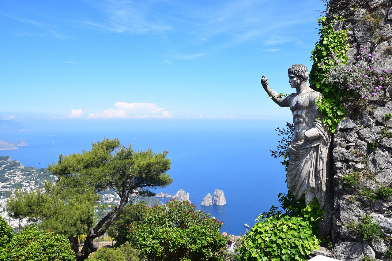 Estatua cl&aacute;sica rodeada de vegetaci&oacute;n con una vista panor&aacute;mica del mar azul y formaciones rocosas en Capri, Italia, ideal para un paquete tur&iacute;stico en 2025.