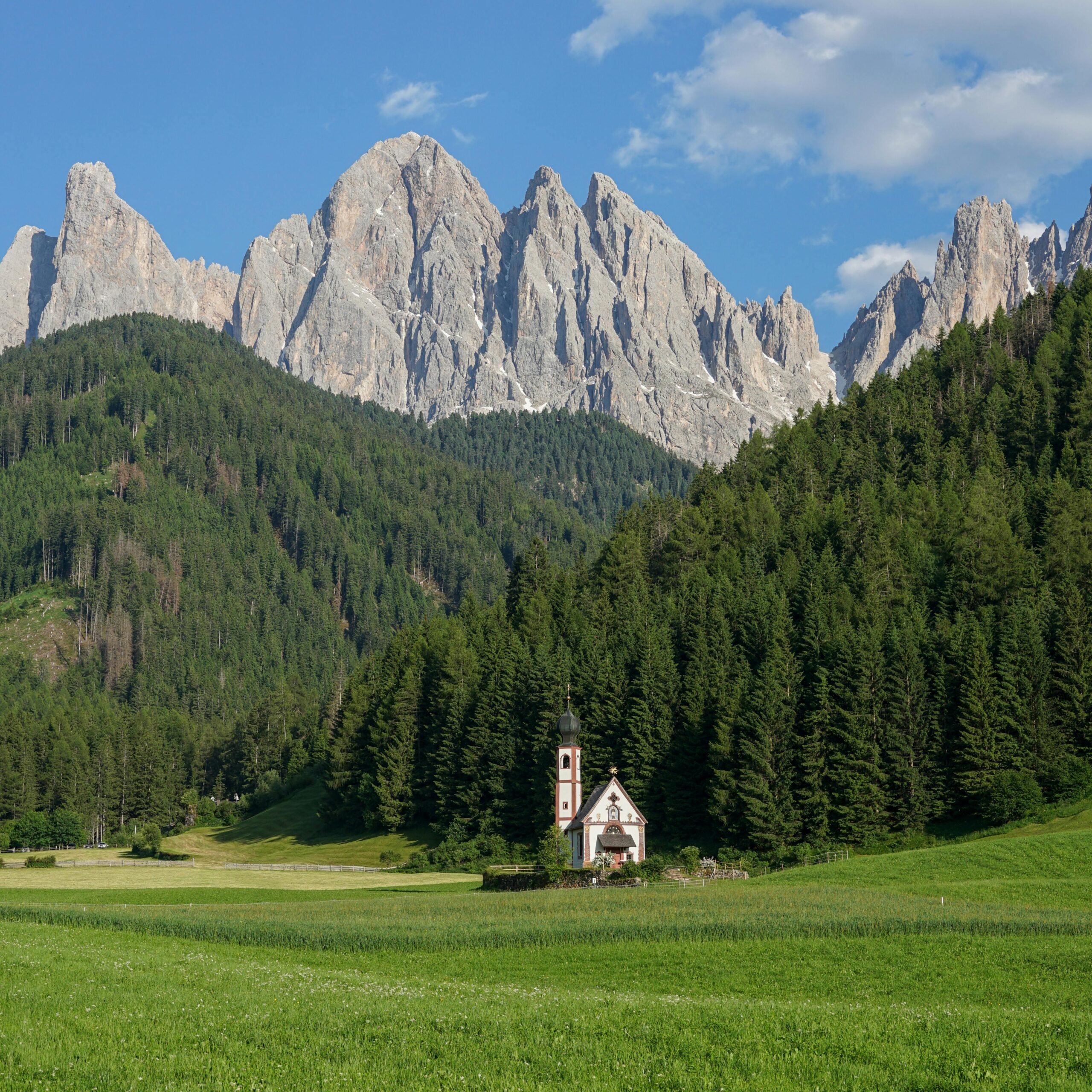 Hermosa vista de una peque&ntilde;a iglesia rodeada de campos verdes y densos bosques de pinos, con los impresionantes picos de las monta&ntilde;as Dolomitas al fondo en un d&iacute;a soleado en Italia.